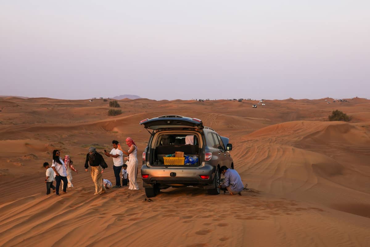Eine Gruppe von Menschen steht neben einem SUV, der auf orangefarbenen Sanddünen in der Wüste von Dubai bei Sonnenuntergang geparkt ist.