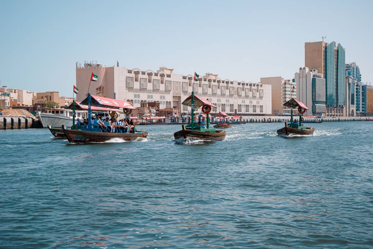 Drei traditionelle Abra-Boote befördern Passagiere über den Dubai Creek, im Hintergrund sind Stadtgebäude und klares blaues Wasser zu sehen.