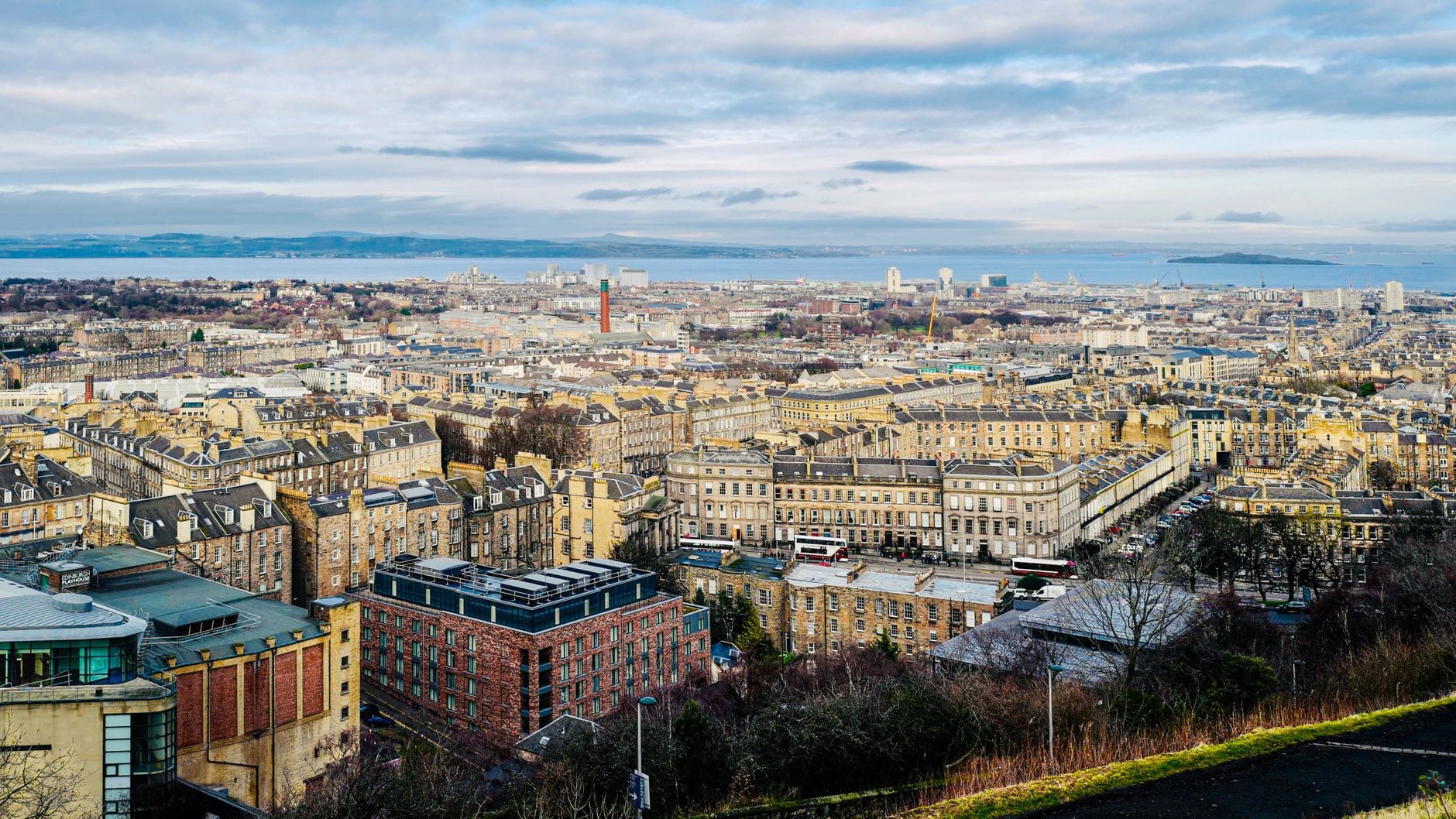 Vista panorámica del paisaje del New Town de Edimburgo con hileras de terrazas georgianas, amplias calles y el Firth of Forth a lo lejos bajo un cielo nublado.