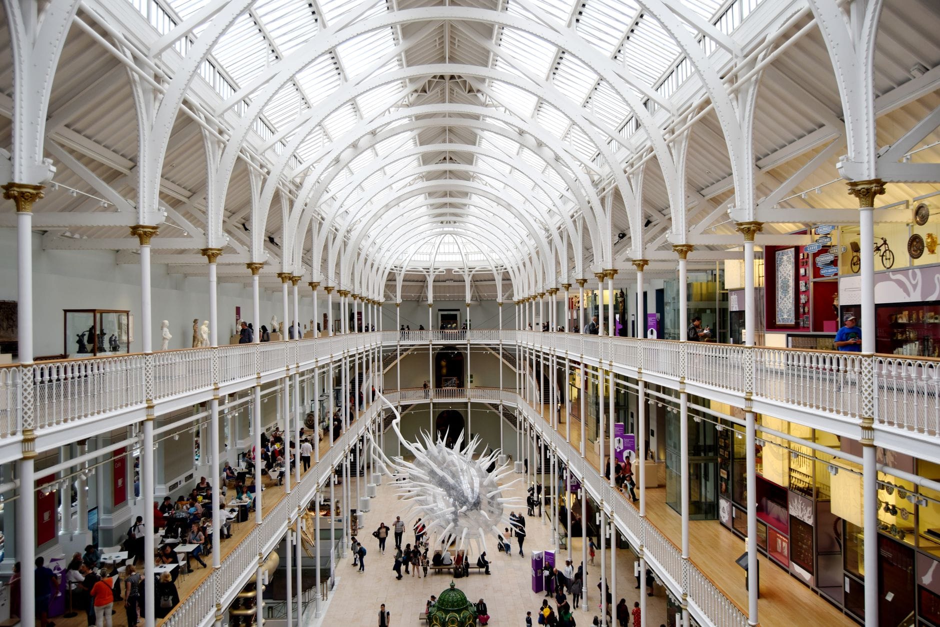Atrio espacioso y luminoso del National Museum of Scotland con visitantes caminando y sentados en bancos, con un dramático techo de hierro forjado blanco.