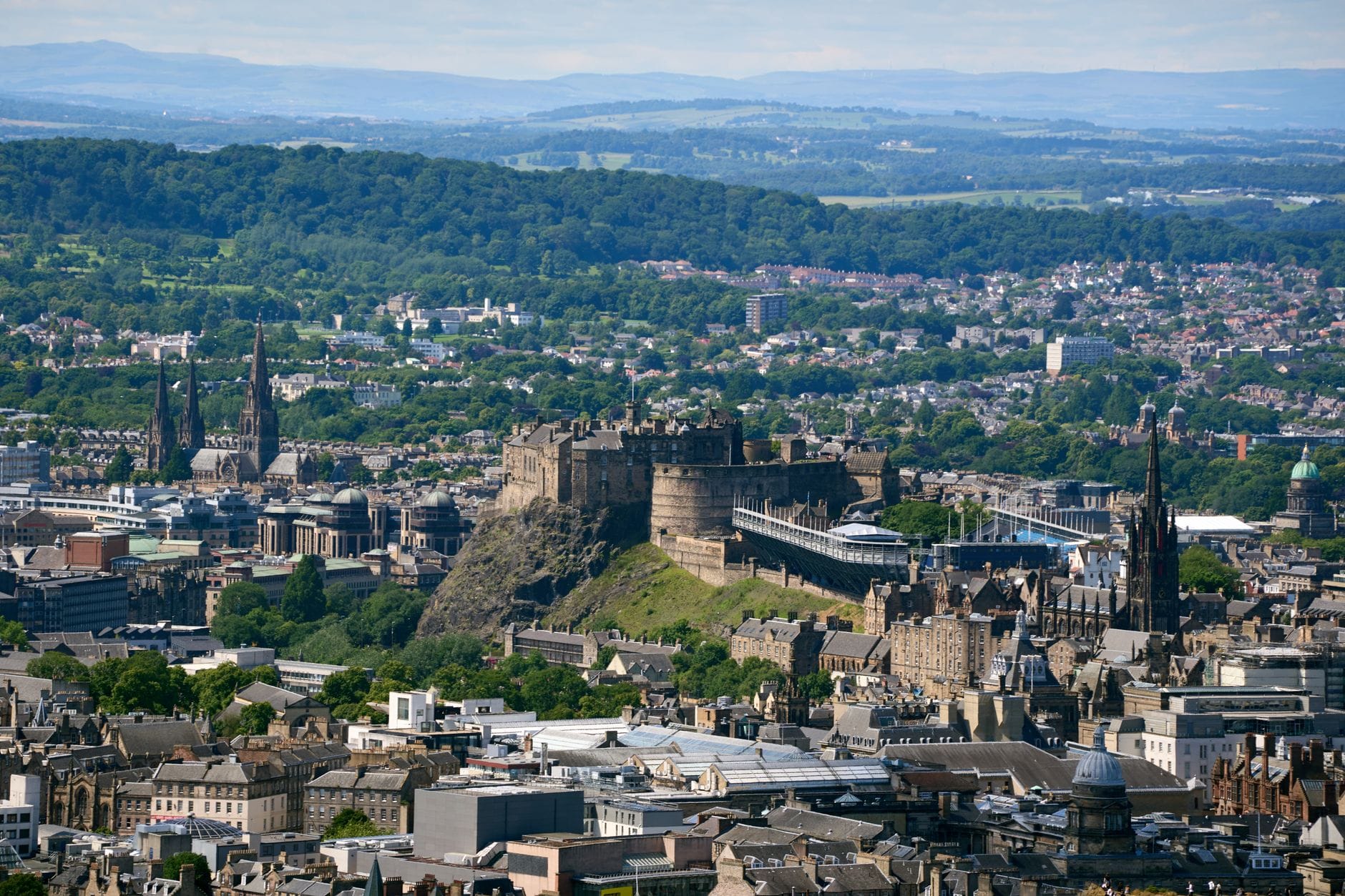 Vista panorámica amplia de Edimburgo mostrando el Castillo de Edinburgh prominentemente en su colina con la ciudad y el verdor extendiéndose a su alrededor.