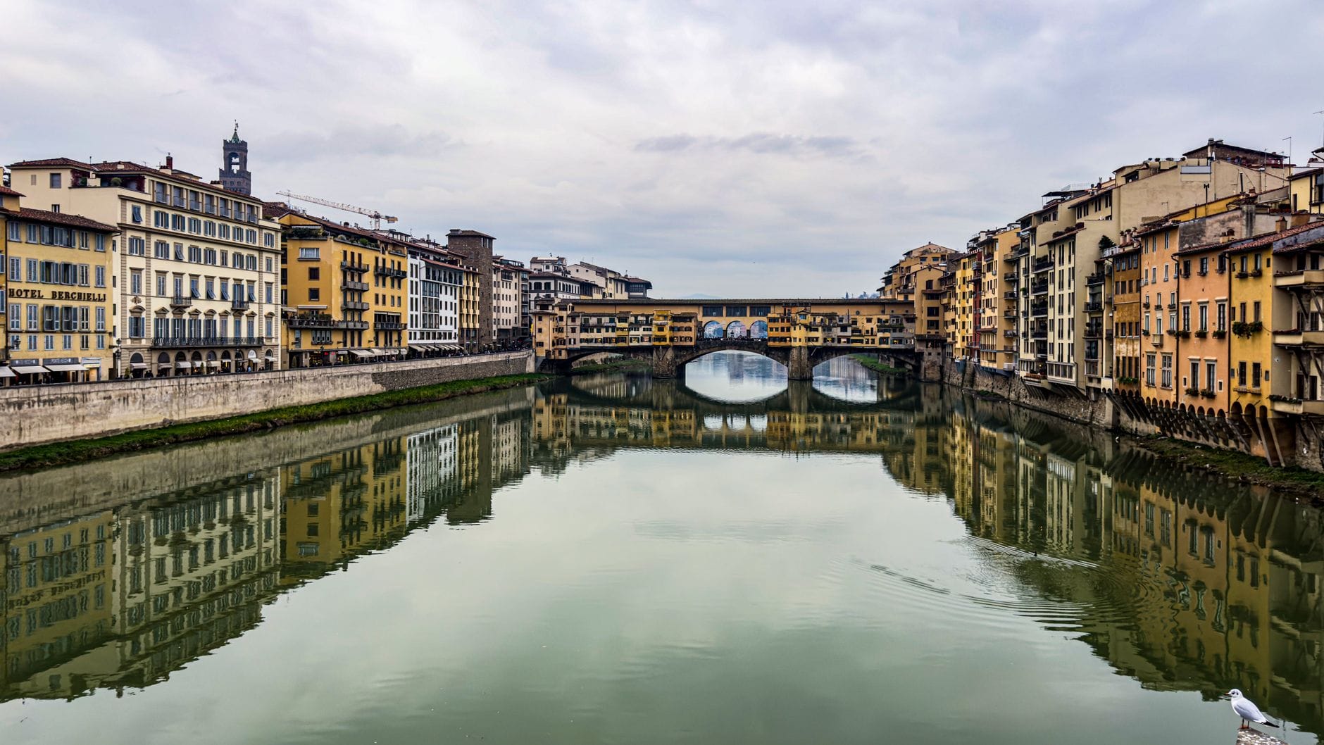 วิวกว้างของสะพาน Ponte Vecchio อันโด่งดังในเมือง Florence ที่ทอดข้ามแม่น้ำ Arno โดยมีอาคารประวัติศาสตร์ขนาบข้างและสะท้อนลงบนผืนน้ำอันสงบ