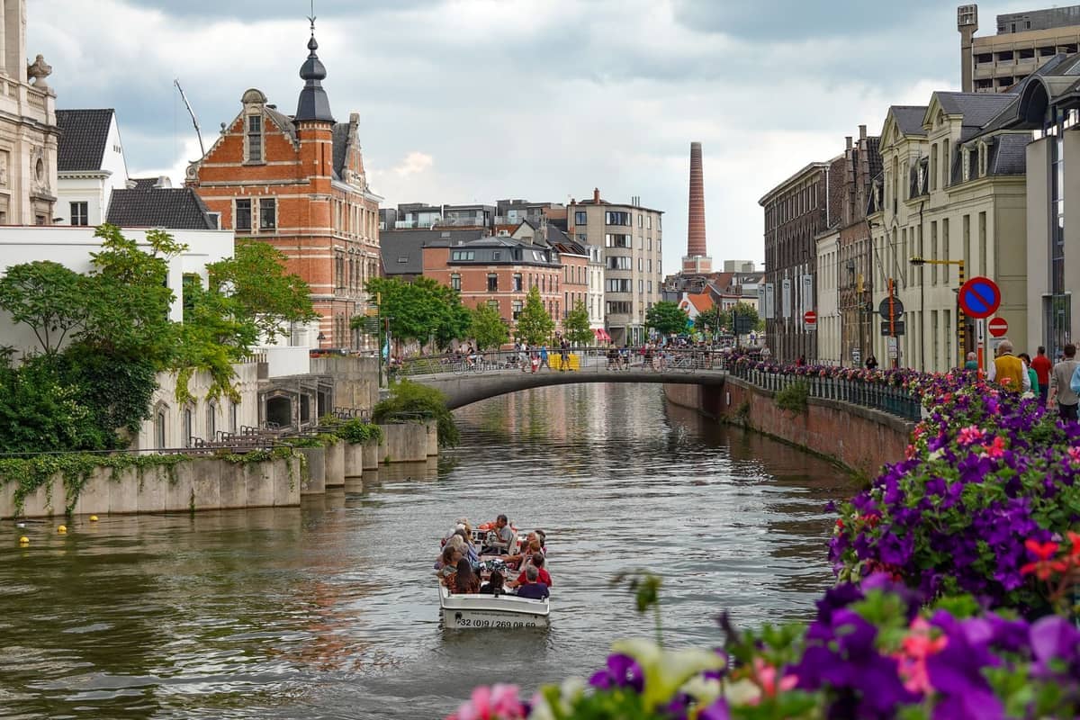 View of a canal in Ghent lined with historic and modern buildings, people boating, and vibrant flowers along the bridge on a cloudy day.