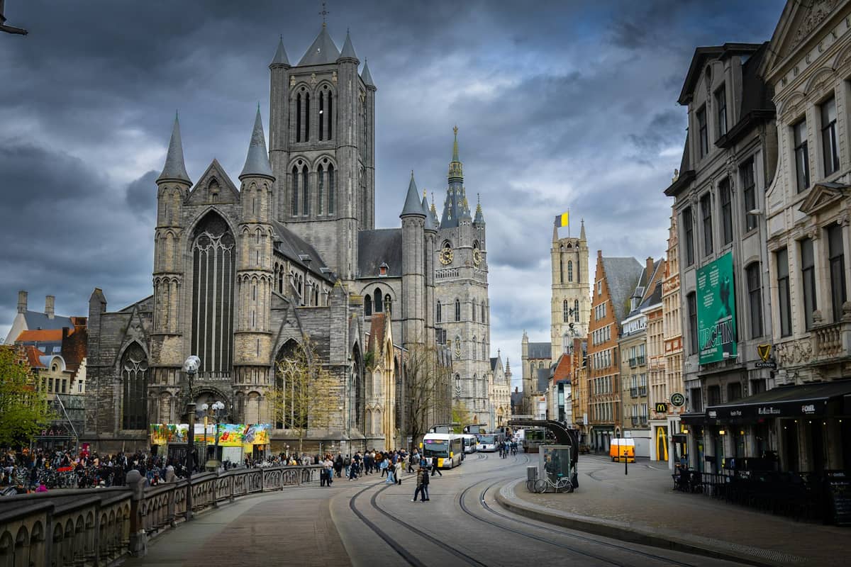 Wide view of Ghent’s historic core showing St Nicholas Church, the Belfry, and St Bavo's Cathedral aligned, with people and tram tracks in front.