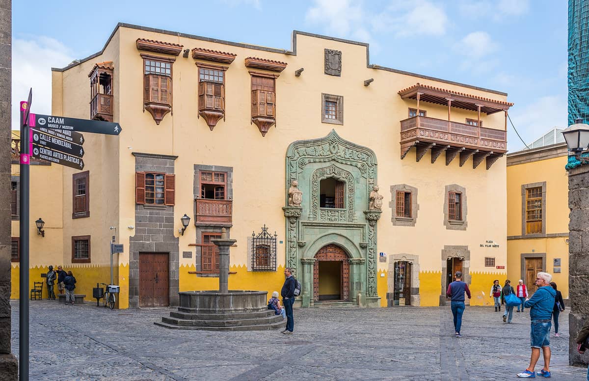 Исторический фасад Casa de Colón с искусной зелёной каменной кладкой и деревянными балконами в старом городе Лас-Пальмас, прохожие и площадь перед зданием.