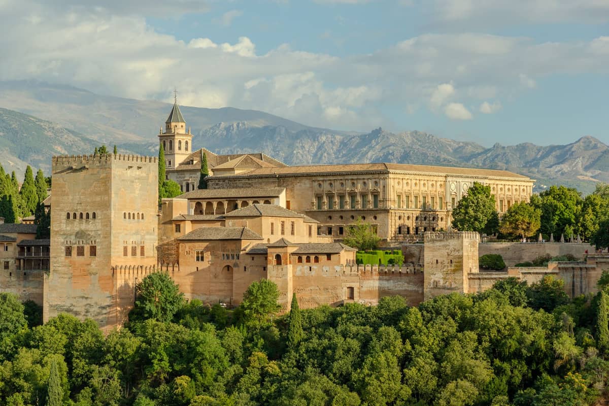 วิวภูมิทัศน์ของพระราชวัง Alhambra ในเมือง Granada พร้อมพืชพรรณเขียวชอุ่มและเทือกเขา Sierra Nevada เป็นฉากหลังภายใต้ท้องฟ้าที่มีเมฆบางส่วน