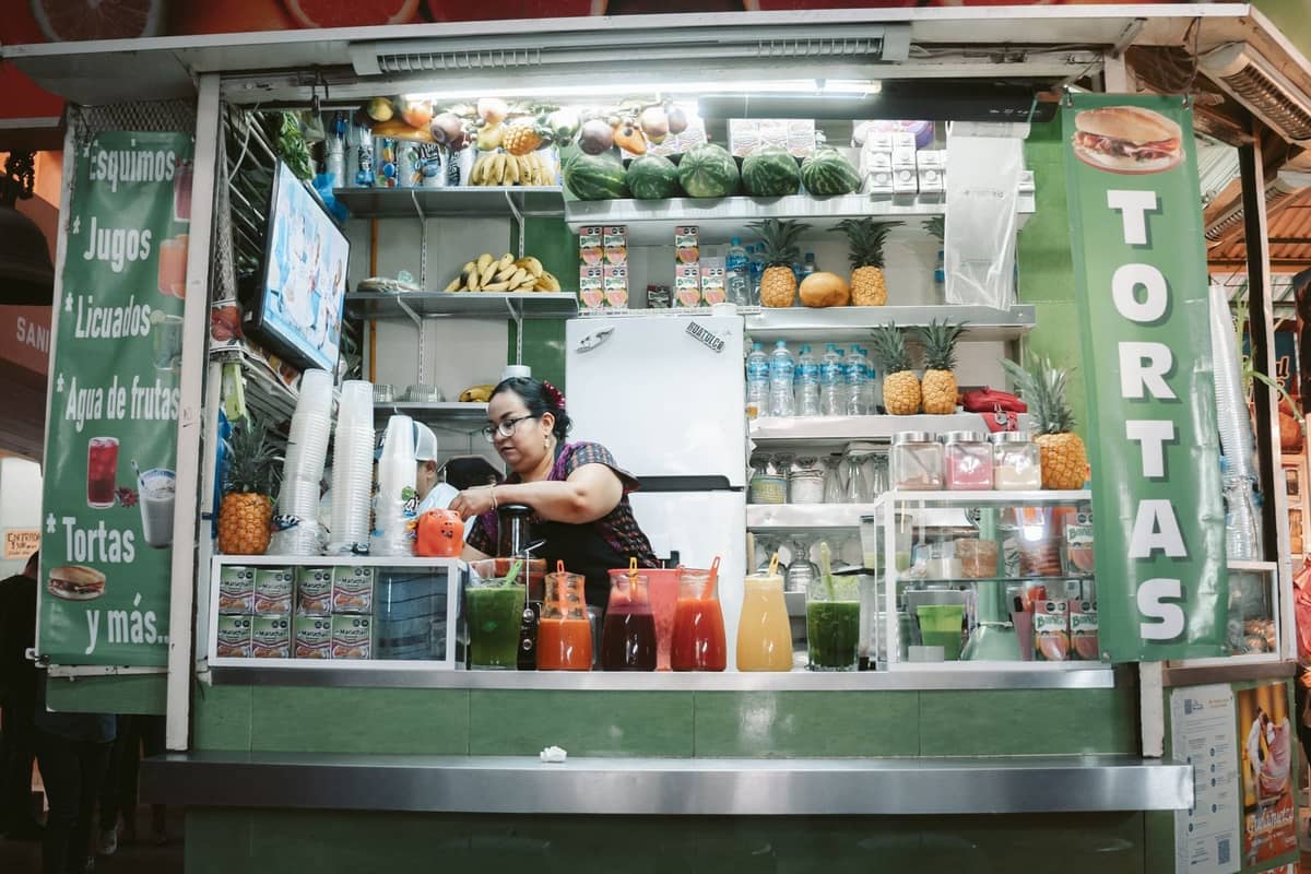Ein Marktimbissstand in Guadalajara, der Tortas, Säfte und frisches Obst verkauft, mit einer Frau, die hinter der Theke Essen zubereitet.