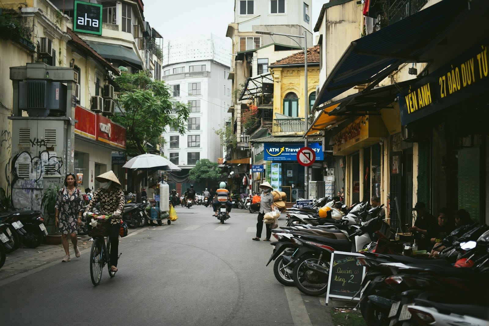 Bustling Hanoi street scene with people walking, riding motorbikes, and sitting at cafes, surrounded by tall yellow colonial-era buildings and shopfronts.