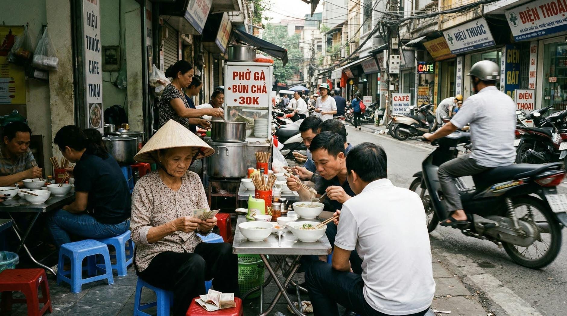 People eating noodle soup at small tables and stools on a busy Hanoi street with street food stalls and signs.