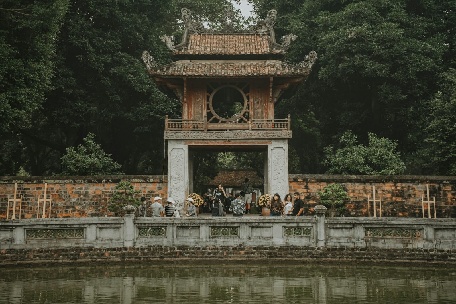 Temple of Literature gate in Hanoi with people and trees reflected in a pond, highlighting a historic and cultural site.