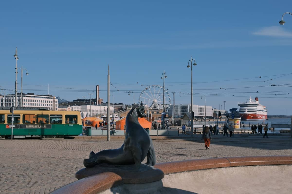 Vista da Praça do Mercado de Helsinki com a estátua da foca, bonde verde, roda-gigante, navio de cruzeiro no porto e pessoas sob um céu azul limpo.