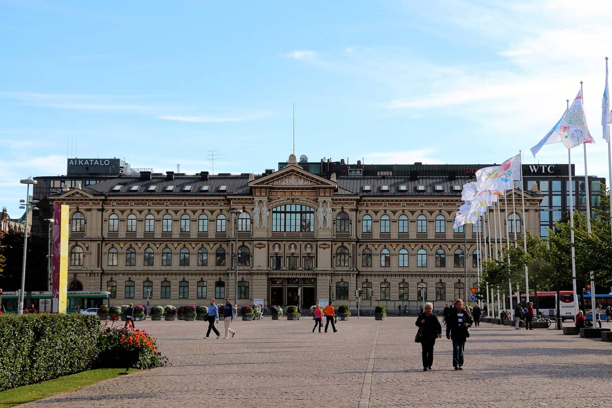 Vista ampla do Ateneum Art Museum em Helsinki com pessoas caminhando pela praça, uma fileira de bandeiras e um céu azul claro.