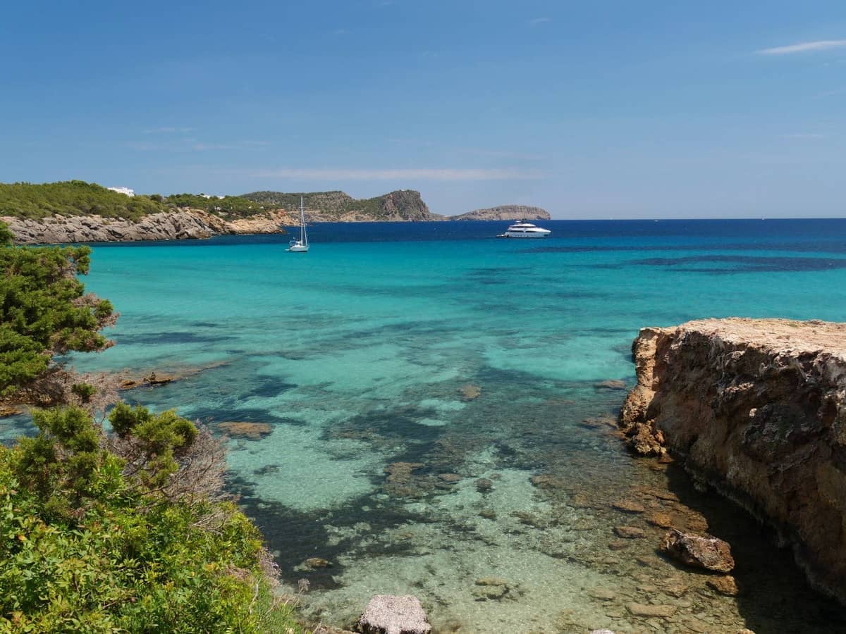 Clear turquoise water and rocky coastline with boats anchored in a picturesque cove. Lush greenery borders the shore under a bright blue sky.