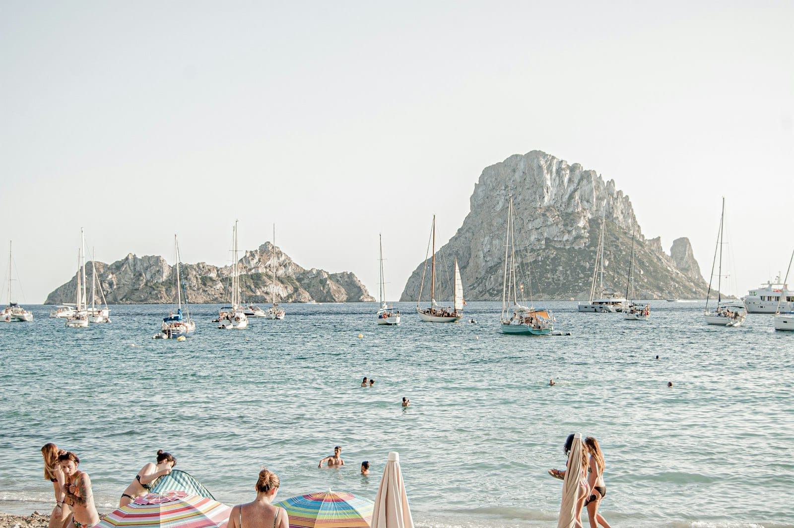 Pessoas relaxando sob guarda-sóis em uma praia de areia de Ibiza, com veleiros ancorados perto da ilha de Es Vedrà e água azul cristalina.