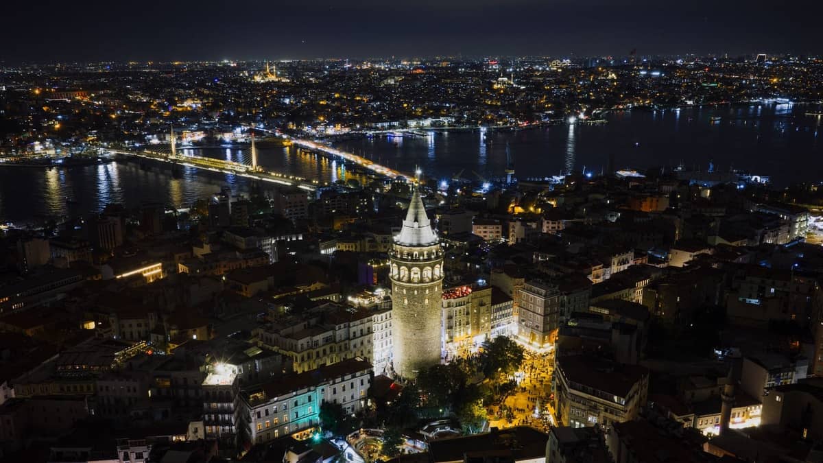 Vue aérienne nocturne d'Istanbul avec la Tour de Galata illuminée, le Bosphore, des ponts éclairés et les lumières de la ville s'étendant au loin.