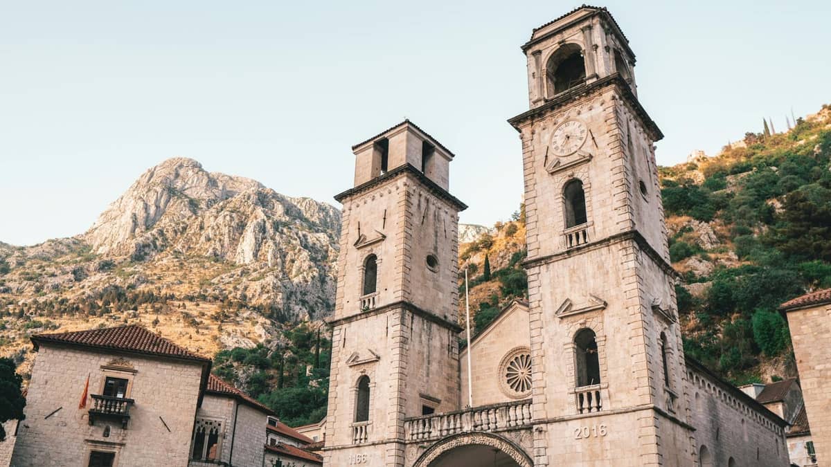 Stone cathedral with two bell towers in Kotor's old town, set against rocky mountains and a clear sky.