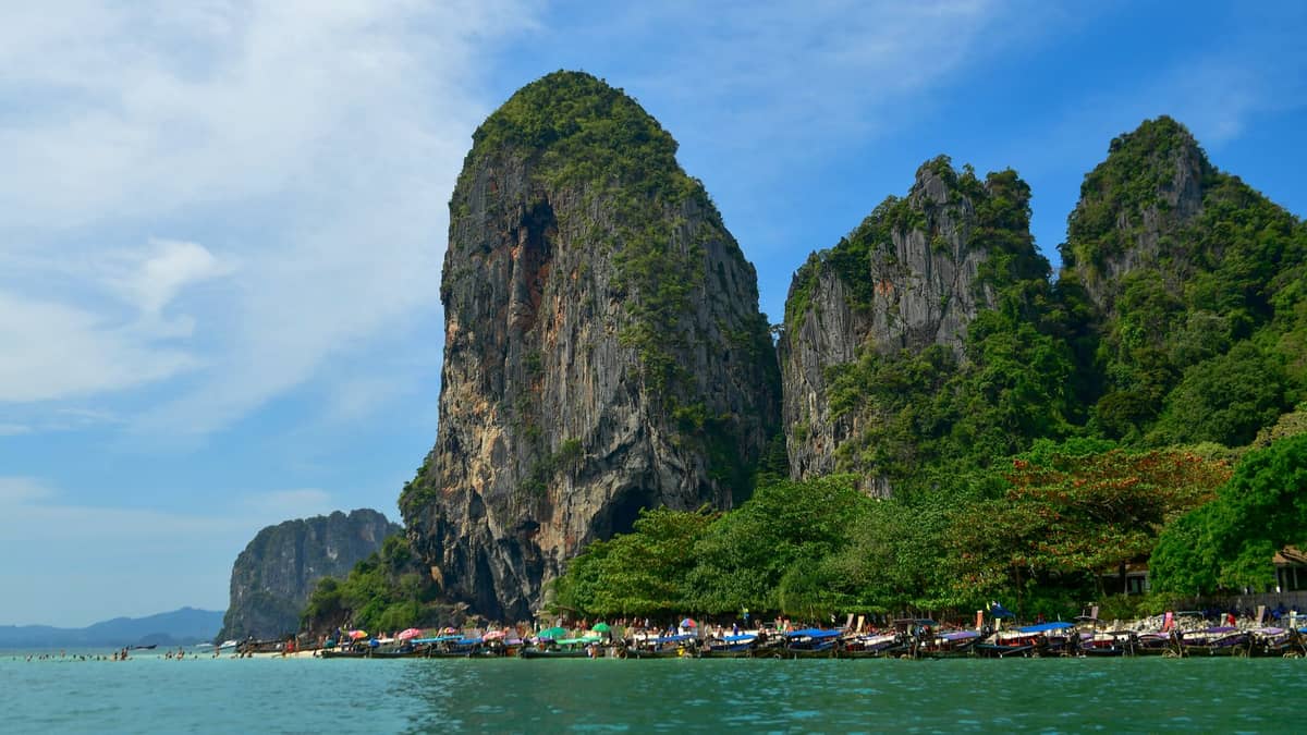 Foto des Railay Beach in Krabi, Thailand, mit türkisfarbenem Wasser, Longtail-Booten entlang der Küste, üppigen grünen Bäumen und aufragenden Kalksteinfelsen.