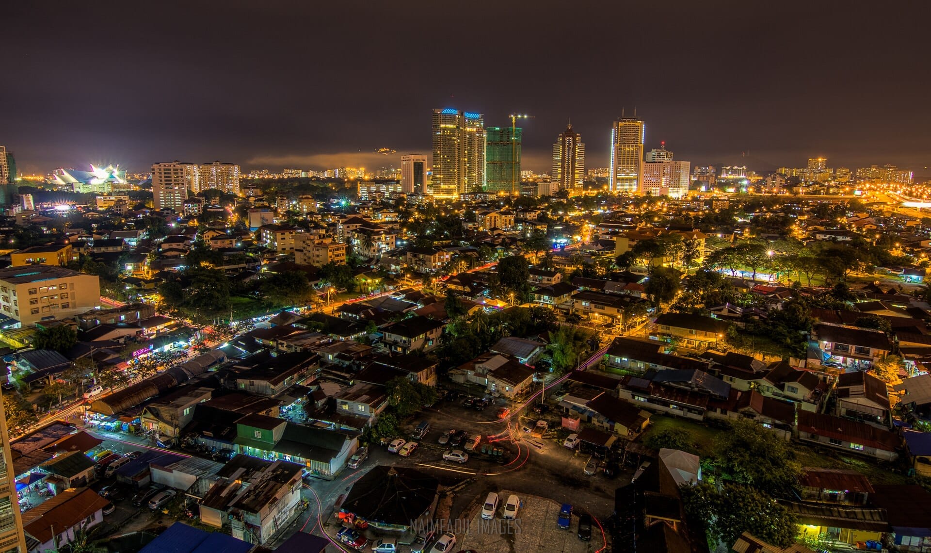 Aerial night view of Kuala Lumpur cityscape with brightly lit skyscrapers, low-rise neighborhoods, and glowing streets under a cloudy sky.