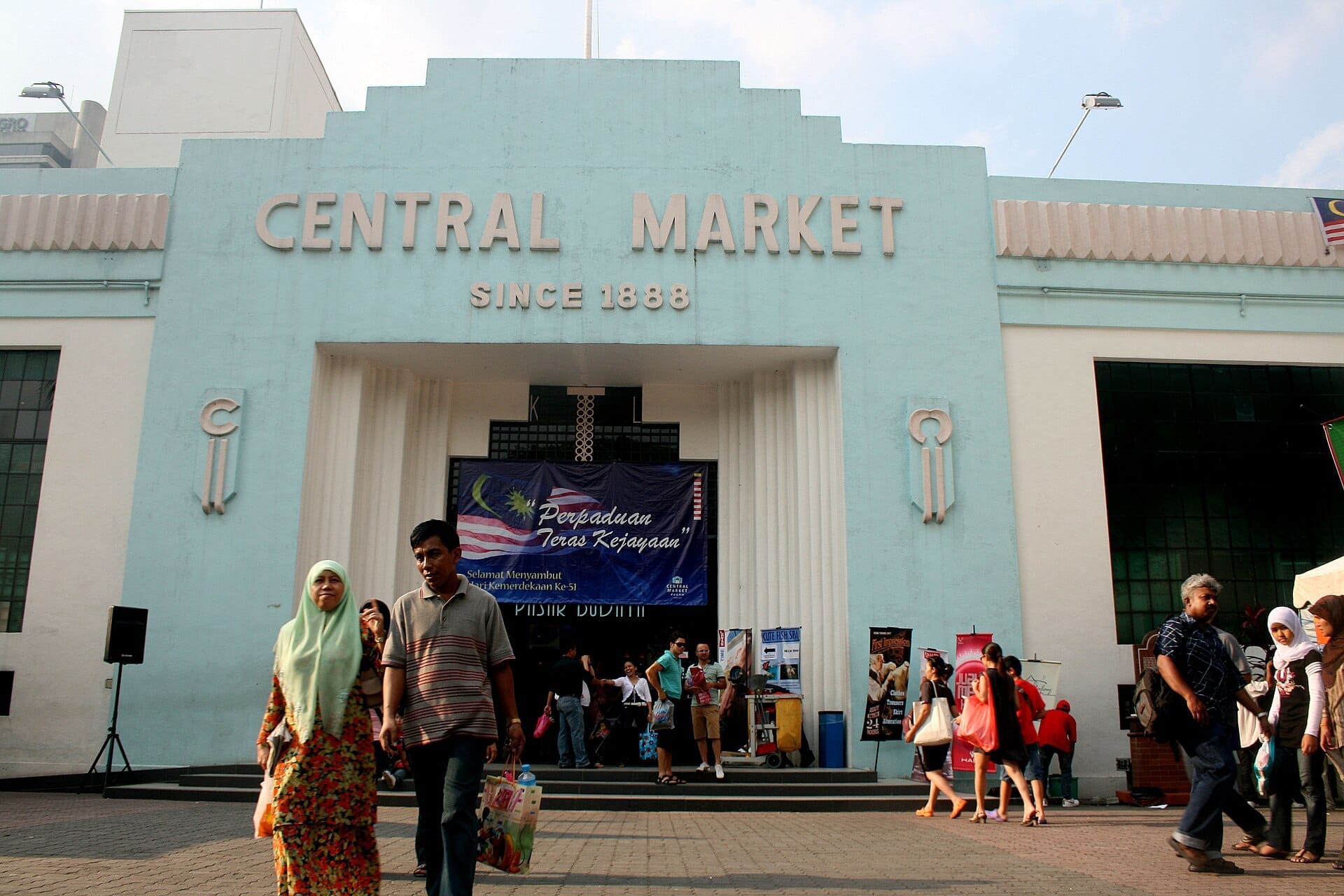 Central Market in Kuala Lumpur with its iconic blue Art Deco facade, people walking by and bright afternoon sunlight overhead.