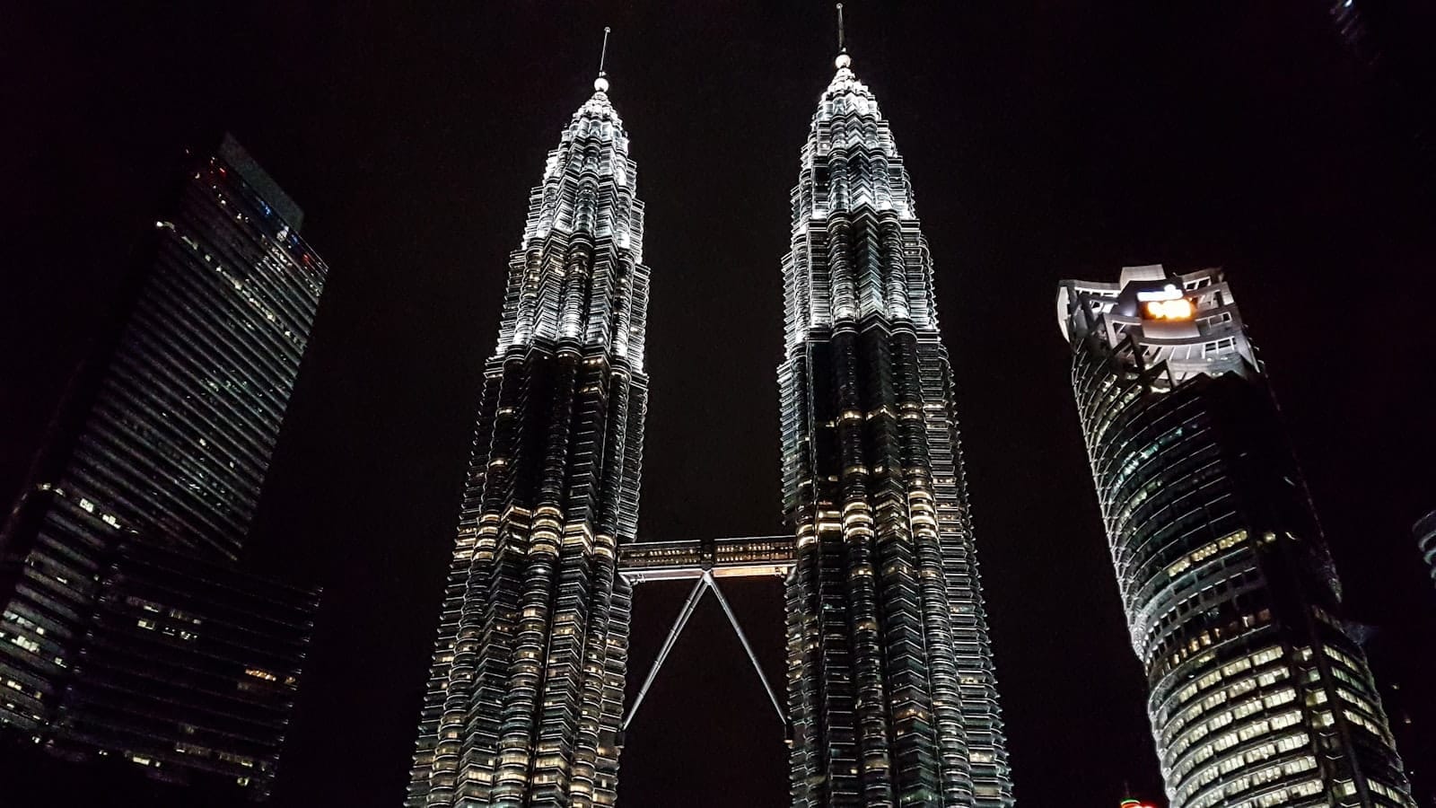 The Petronas Twin Towers in Kuala Lumpur illuminated at night, flanked by other modern high-rise buildings against a dark sky.