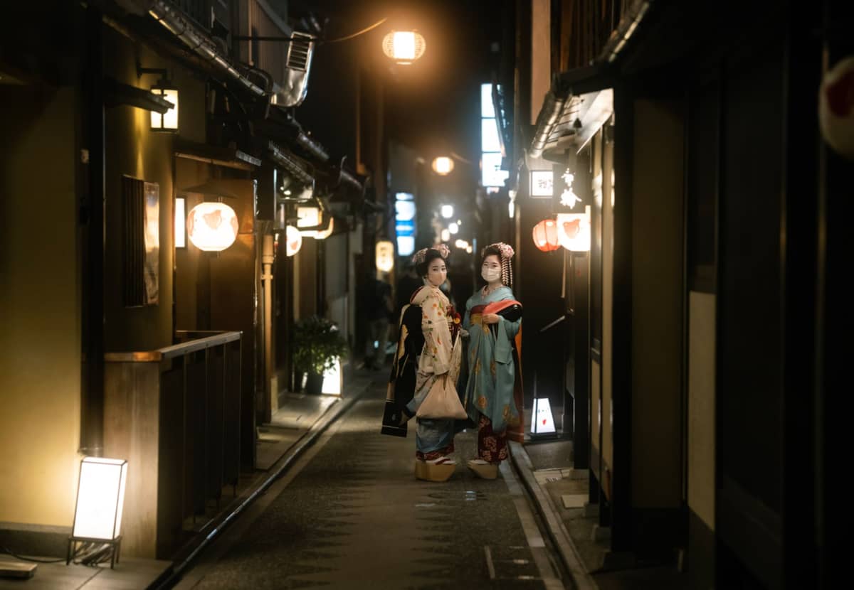 Two women in kimono with masks walking a narrow lantern-lit alley in Kyoto’s historic district at night, lined with wooden buildings.