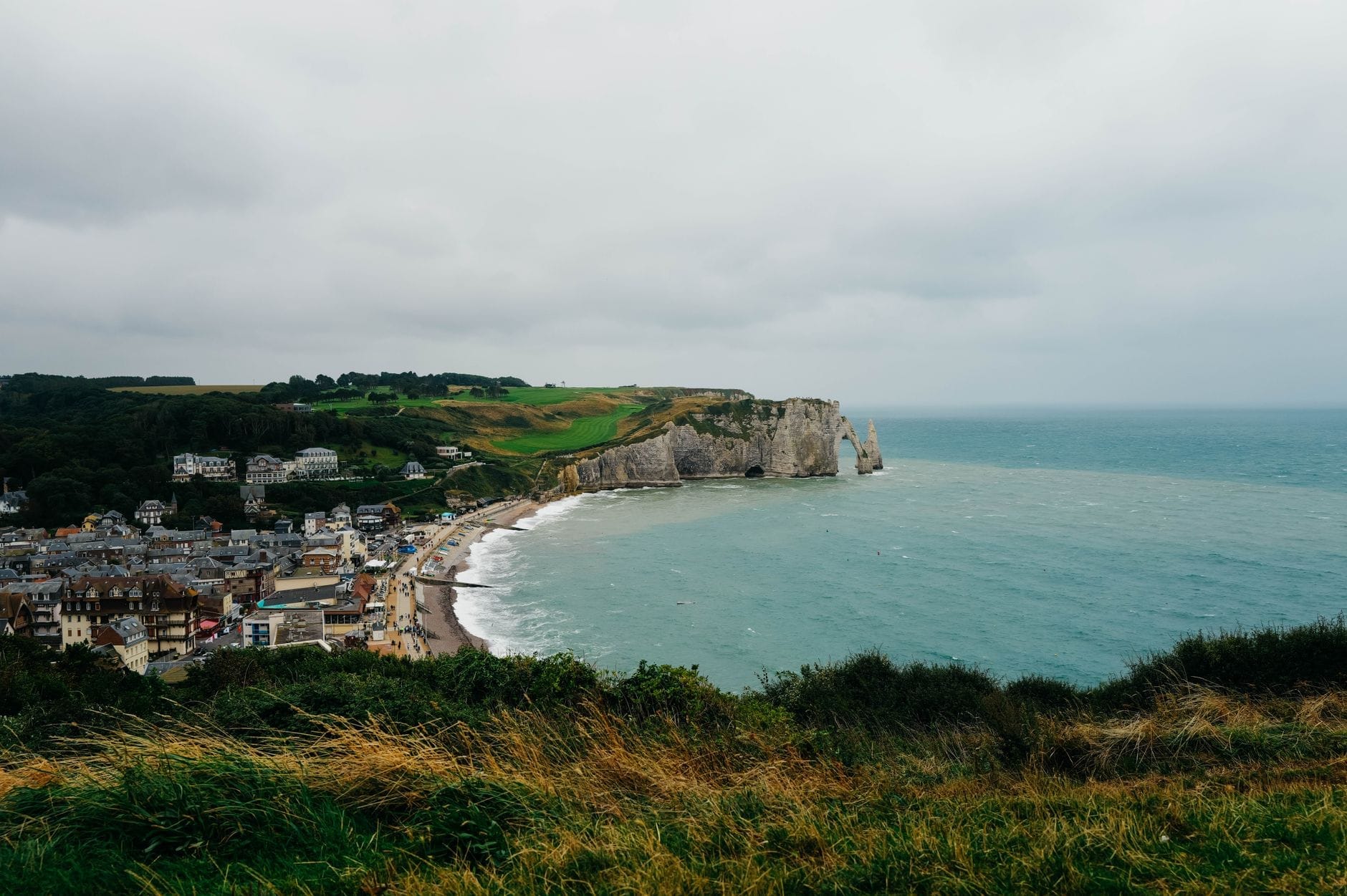 Panoramiczny widok na wioskę Étretat i jej kredowe klify na wybrzeżu Normandii pod zachmurzonym niebem, widziany z wysokiego punktu widokowego.