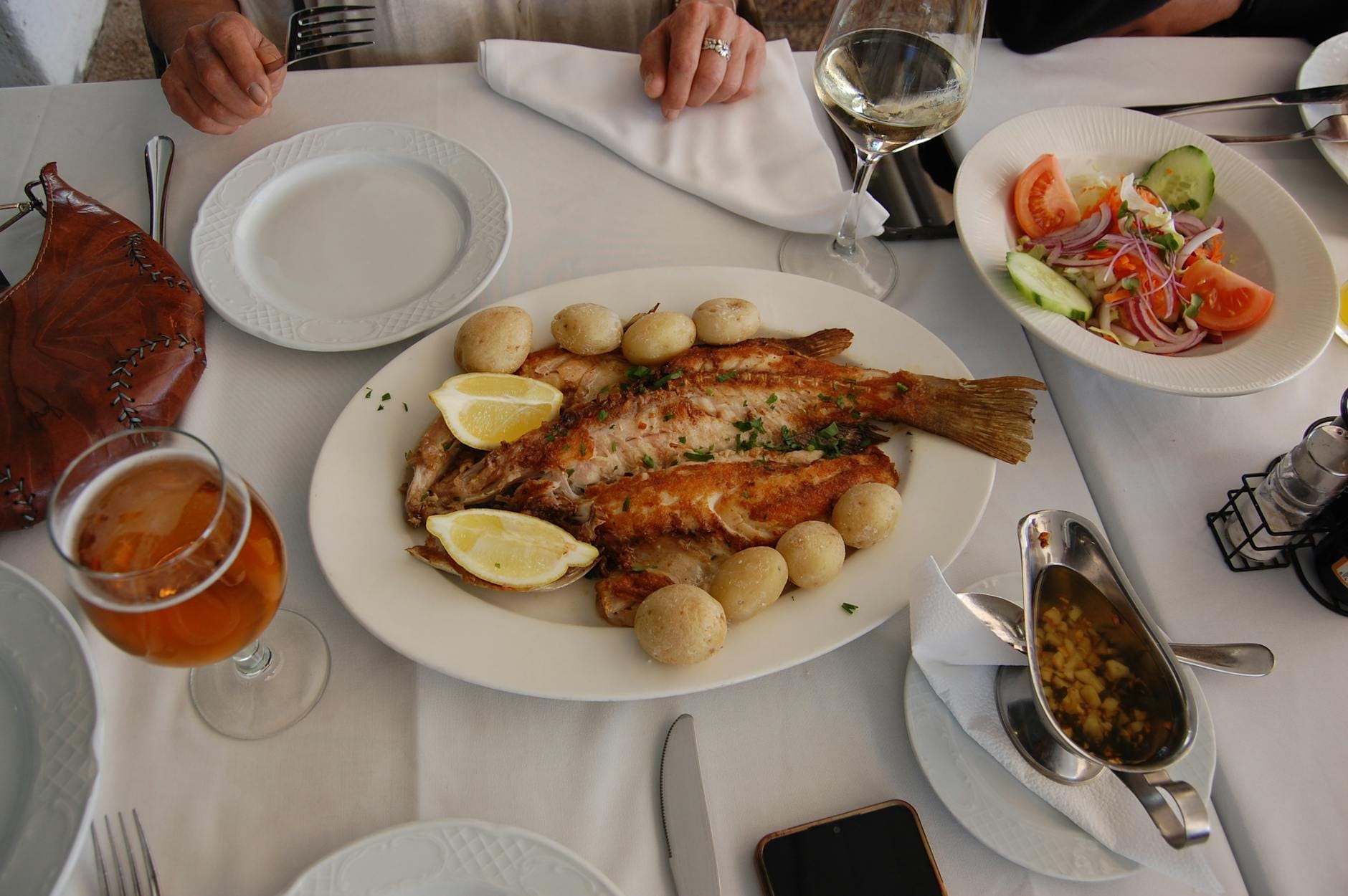 Une table dressée avec des assiettes de poisson grillé, pommes de terre, salade, vin et bière ; les mains des convives sont visibles, évoquant un repas dans un restaurant portugais.