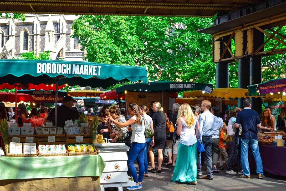 Multidões a explorar bancas de comida sob coberturas verdes com a inscrição Borough Market num ambiente animado ao ar livre com produtos e mercadorias visíveis.