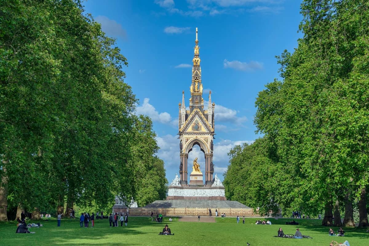 Pessoas relaxando e caminhando na grama em frente ao ornamentado Albert Memorial nos Kensington Gardens de Londres, rodeado de exuberantes árvores verdes.