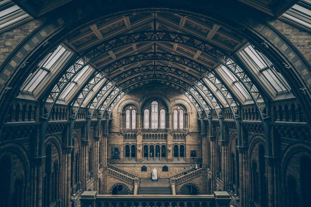 Vista ampla do grande salão com arcos no interior do Natural History Museum de Londres, com decoração em pedra e pessoas na varanda.