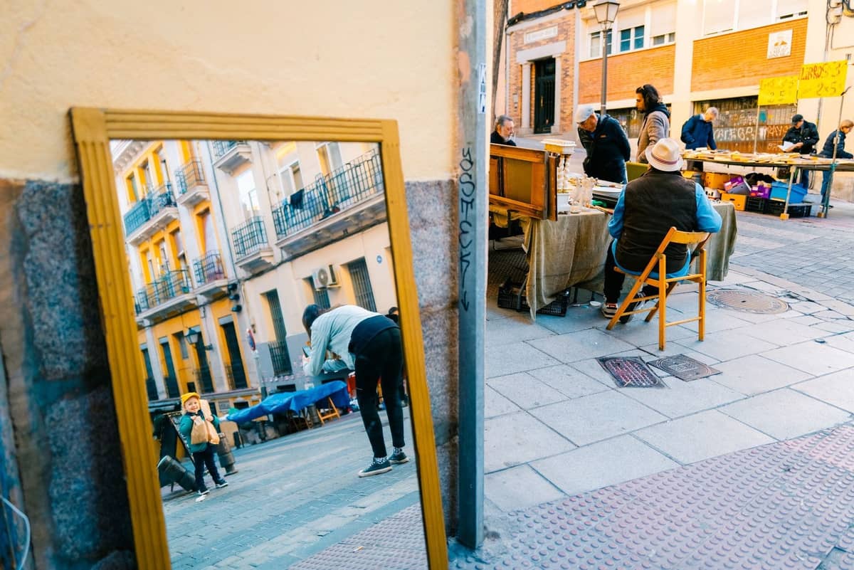 Colorido mercado callejero en Madrid con gente curioseando en los puestos, un espejo que refleja la vida urbana y edificios históricos del barrio.