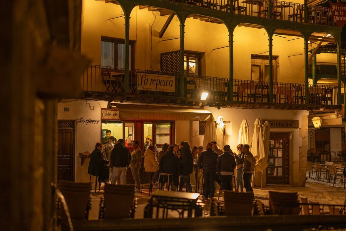 Personas reunidas en el exterior de un bar con luz cálida en una plaza tradicional de Madrid por la noche, evocando la vibrante vida nocturna de la ciudad.