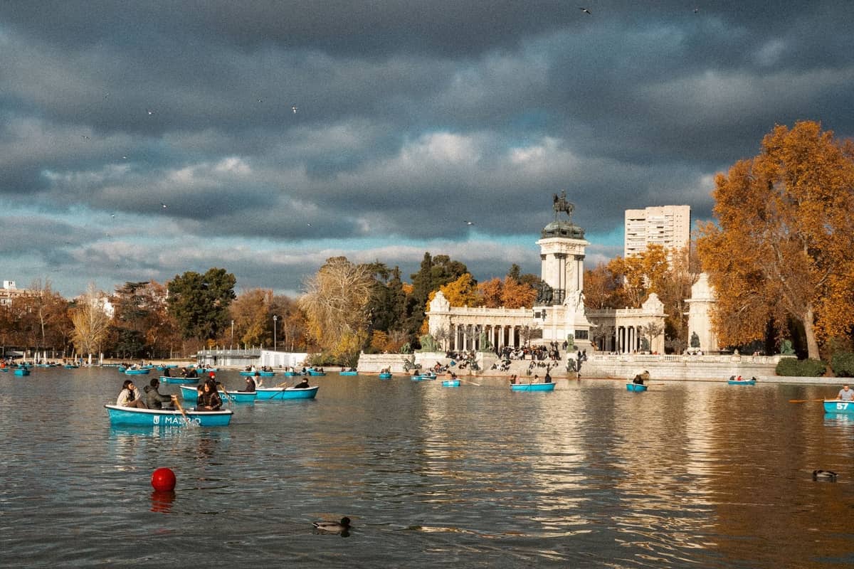 Personas remando en botes azules en el gran estanque frente al Monumento a Alfonso XII en el Parque del Retiro de Madrid en un día nublado.