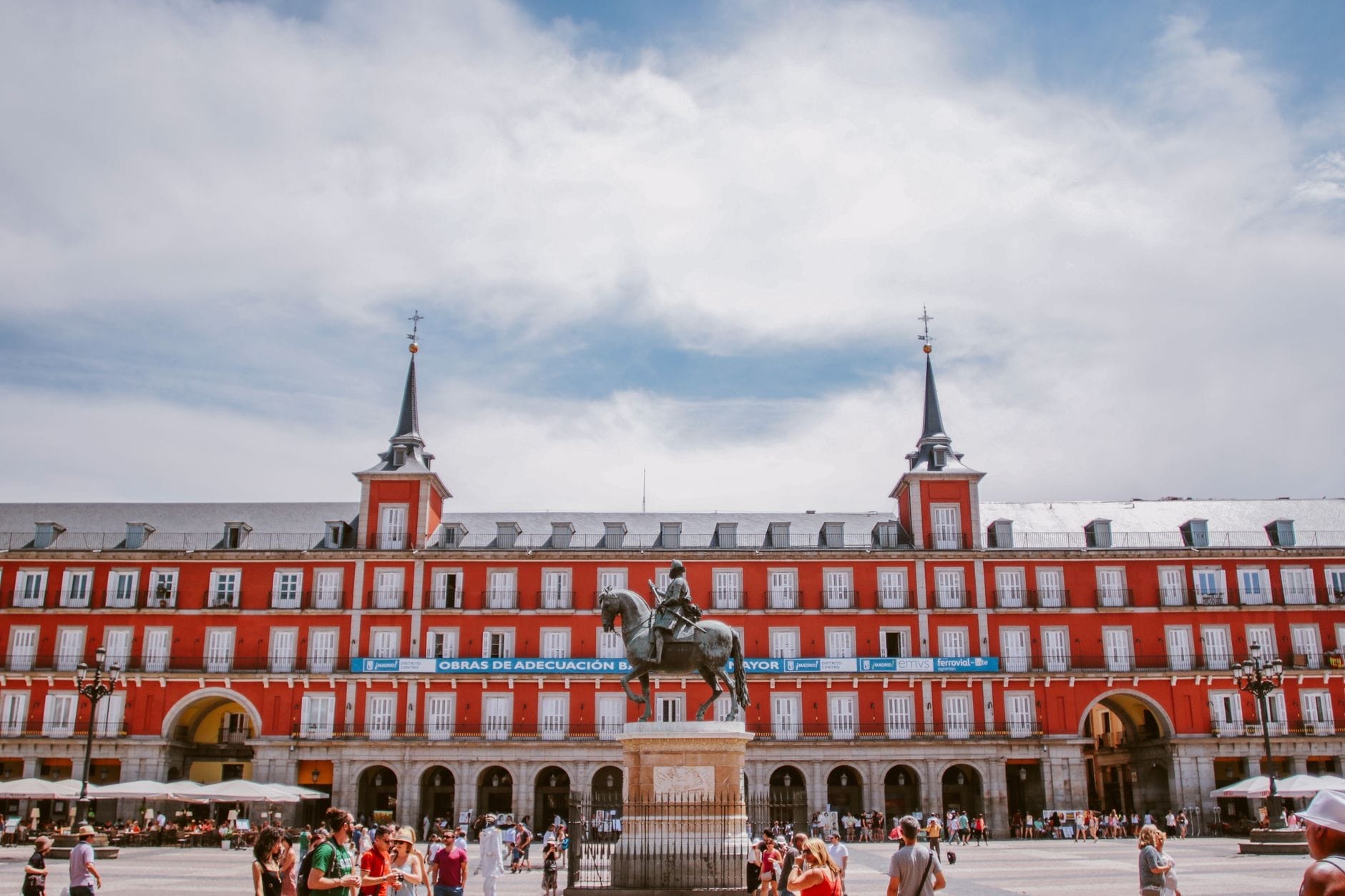 Plaza Mayor em Madri com pessoas caminhando e um céu azul limpo, com os icônicos prédios vermelhos e a estátua equestre ao centro.