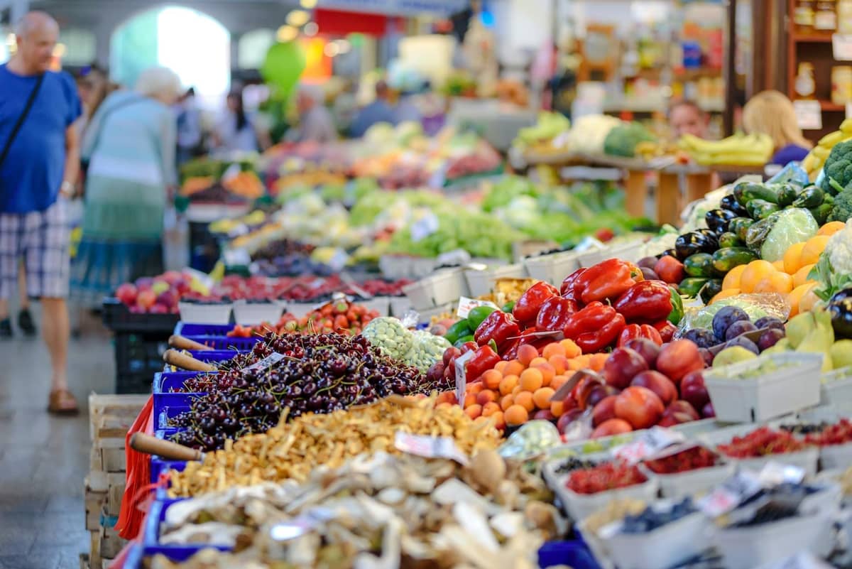 Vibrant indoor market with stalls full of fresh fruits and vegetables, shoppers browsing, and a lively, colorful atmosphere.