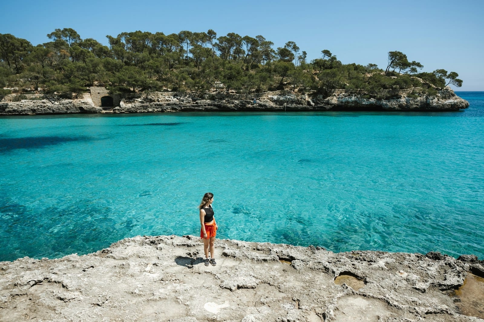 Mulher em pé numa costa rochosa com vista para as águas turquesas cristalinas de Cala Mondragó em Maiorca, com falésias cobertas de pinheiros ao fundo.