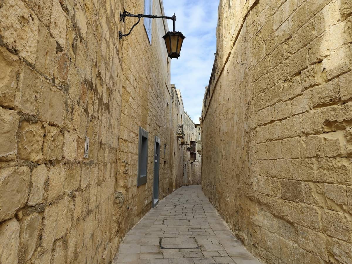 Enge Steingasse mit hohen goldenen Kalksteinmauern und einer hängenden Laterne, typisch für Mdinas ruhige historische Straßen.