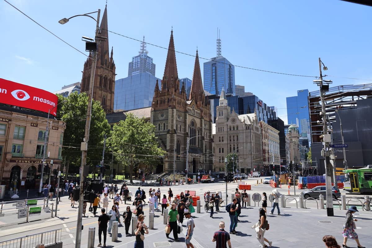 Federation Square mit der St Paul's Cathedral und den Wolkenkratzern von Melbourne, belebt mit Menschen und Straßenbahnen an einem sonnigen Tag