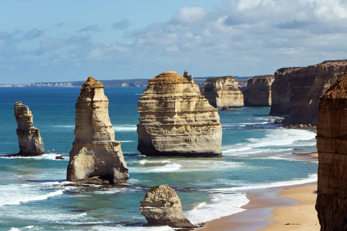 Mehrere Kalksteinformationen, bekannt als die Twelve Apostles, ragen entlang einer dramatischen Küstenlinie mit Wellen und goldenem Sandstrand aus dem Meer.