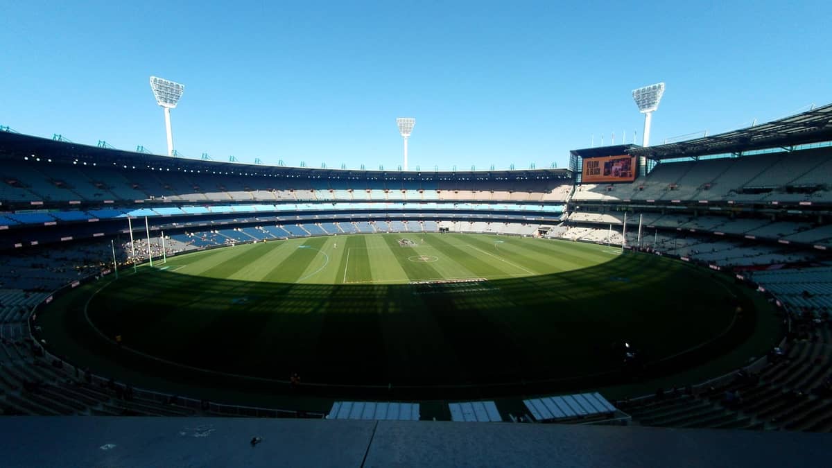 Weitwinkel-Landschaftsansicht des Melbourne Cricket Ground mit leeren Sitzen, grünem ovalem Spielfeld und hohen Stadionlichtern unter einem klaren blauen Himmel.