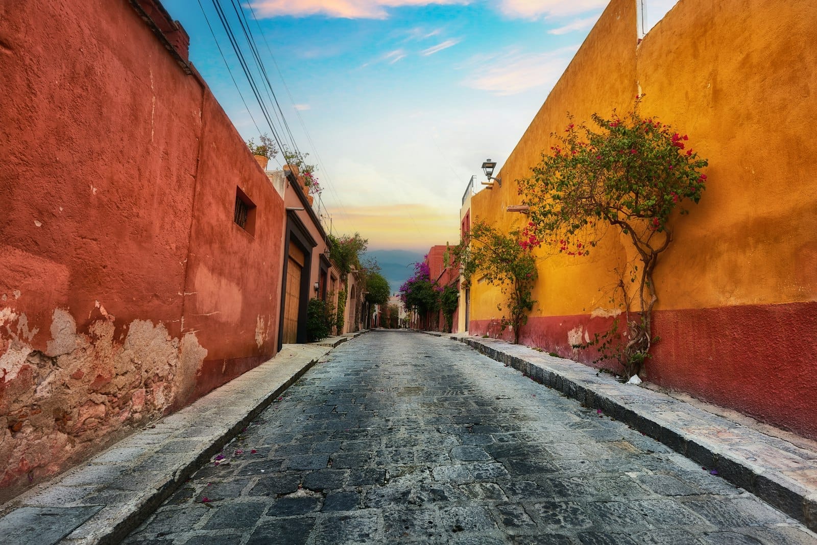 Rua de pedra calçada ladeada por vibrantes paredes vermelhas e amarelas, com uma árvore e um suave céu ao pôr do sol.