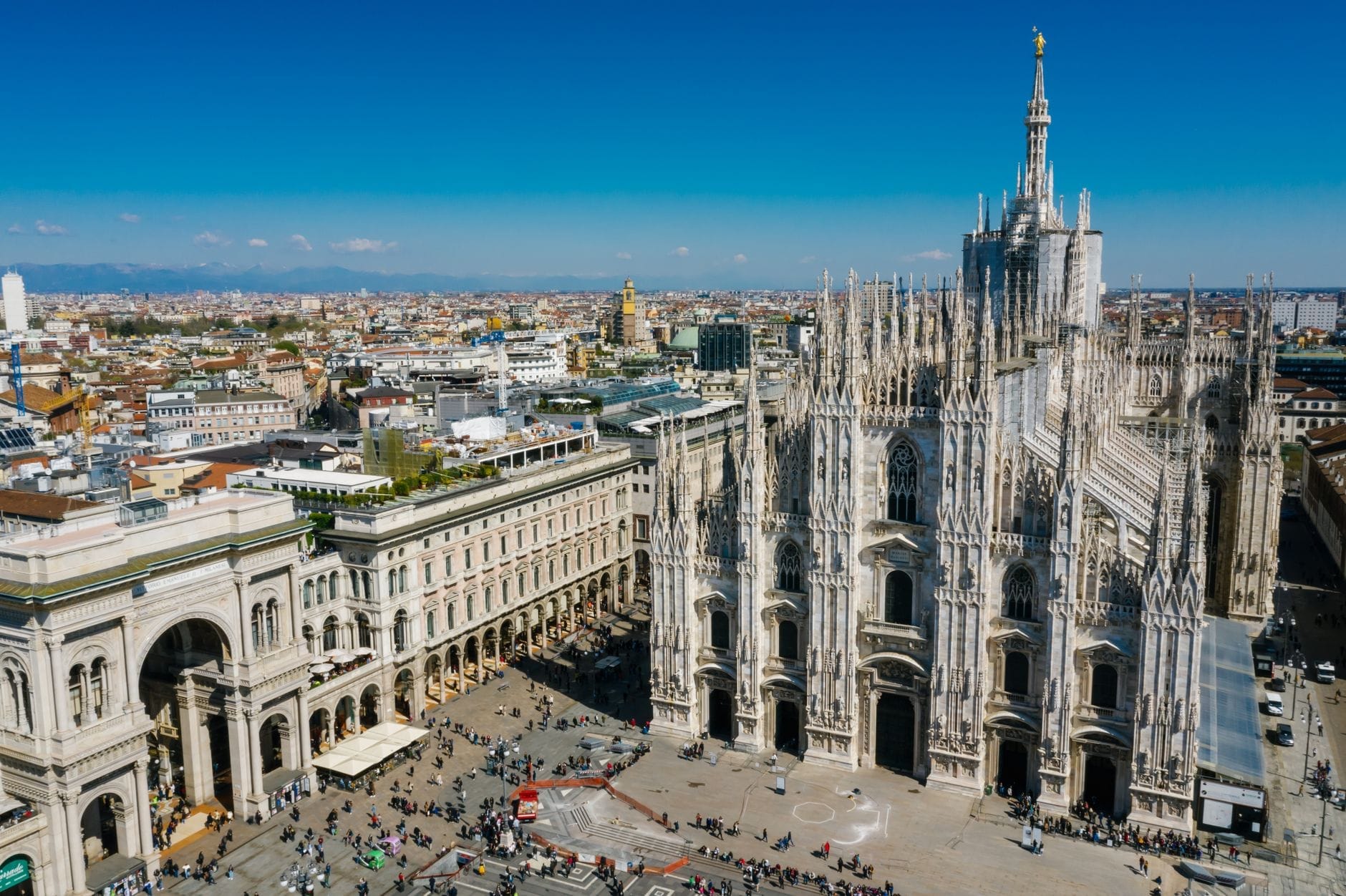 Vista aérea deslumbrante da catedral Duomo de Milão e da Galleria Vittorio Emanuele II, com multidões movimentadas e a paisagem urbana sob um céu azul límpido.