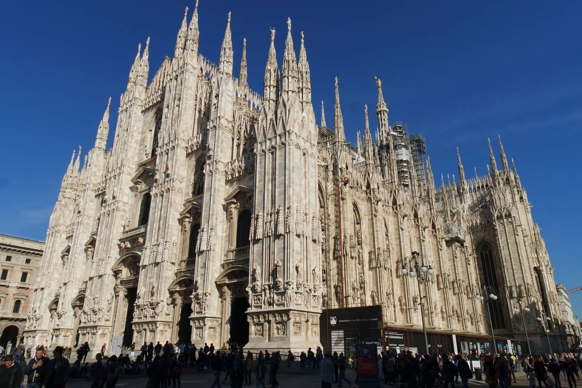 Vista ampla da catedral Duomo de Milão com suas elaboradas torres, fachada de mármore e multidões em primeiro plano sob um céu azul claro.