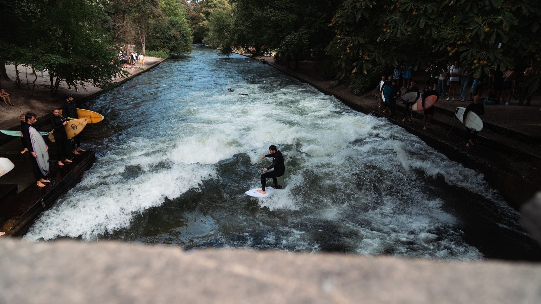 วิวนักเซิร์ฟที่กำลังเล่นคลื่นของแม่น้ำ Eisbach ใน Englischer Garten เมือง Munich โดยมีผู้คนชมอยู่ทั้งสองฝั่งแม่น้ำท่ามกลางพื้นที่สีเขียว