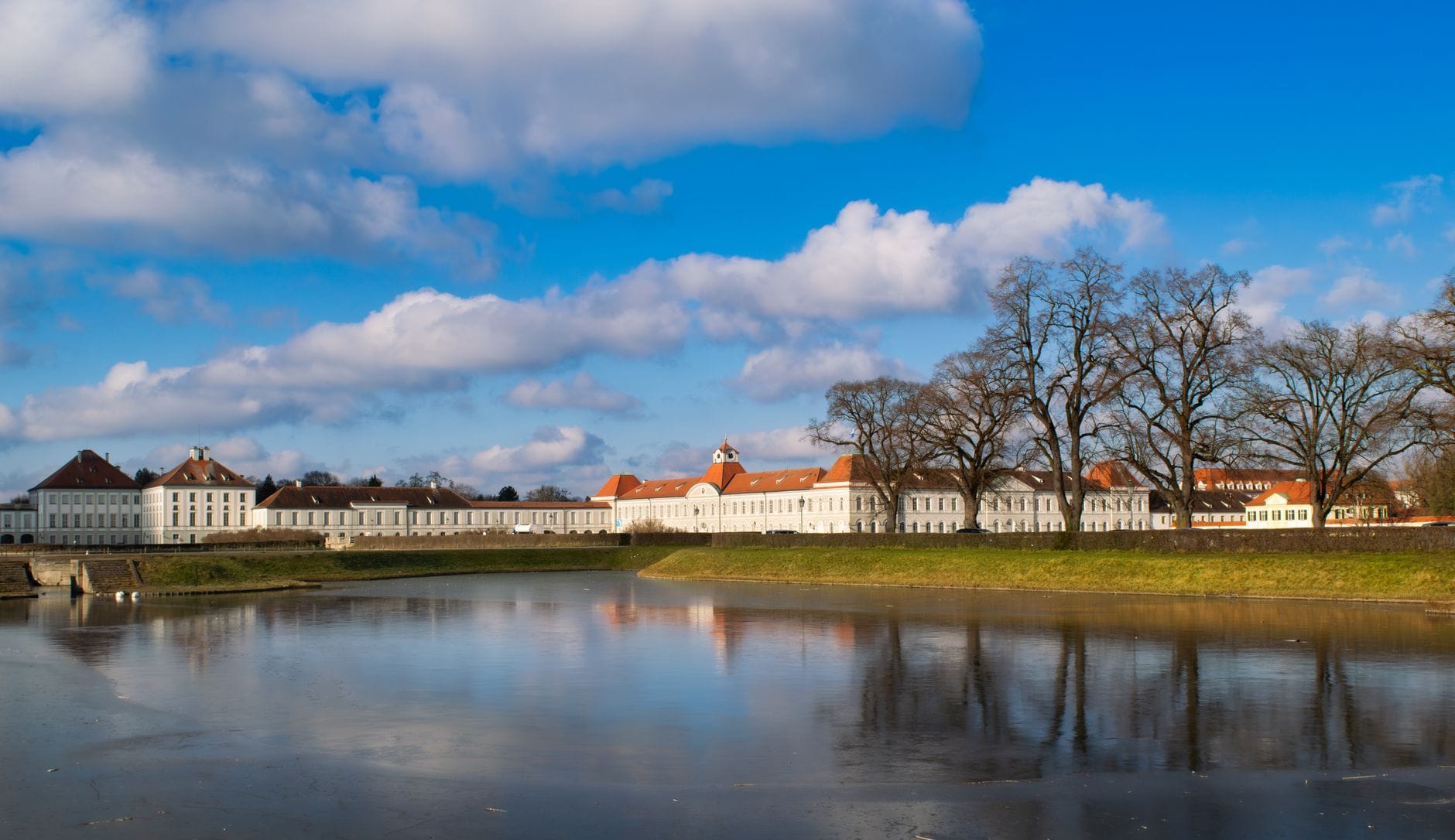 วิวกว้างของพระราชวัง Nymphenburg พร้อมด้านหน้าสไตล์บาโรกที่สะท้อนในบึงน้ำ ต้นไม้ และท้องฟ้าสีฟ้าในเมือง Munich