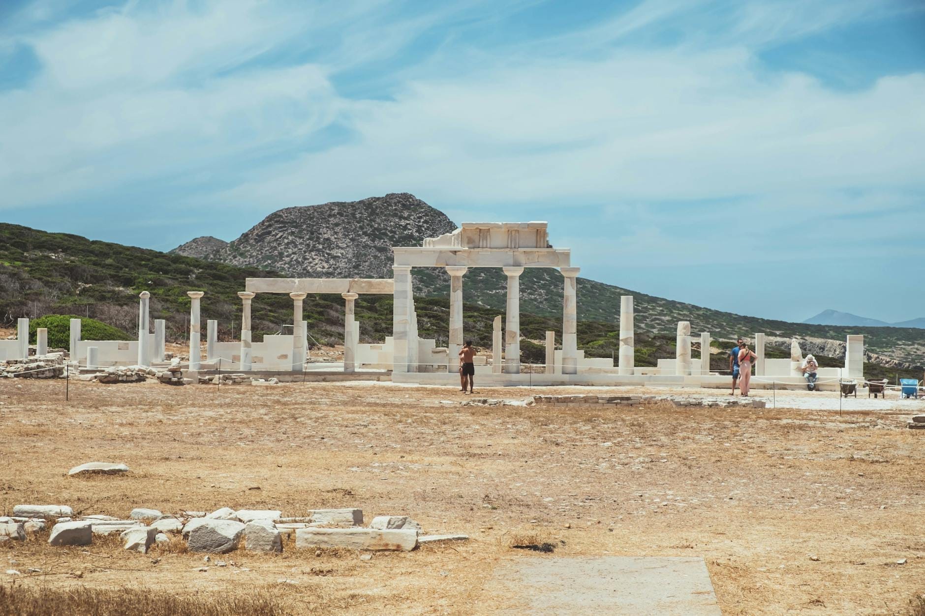 Ruinas antiguas con columnas y turistas en el sitio arqueológico de Delos bajo un cielo azul con montañas al fondo.