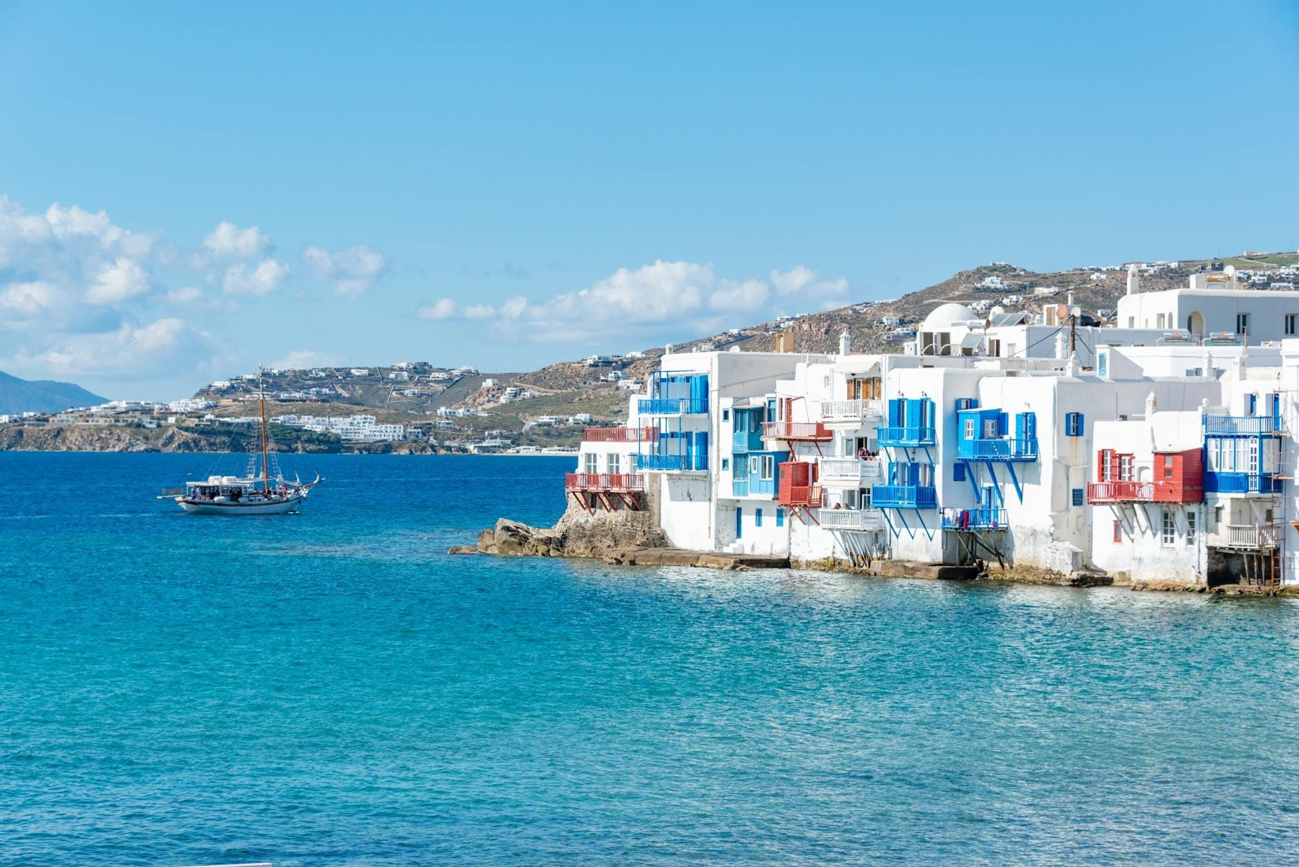 Mar azul brillante con las icónicas casas blancas cicládicas y balcones coloridos del paseo marítimo de Mykonos, cielo despejado y colinas al fondo bajo el sol.