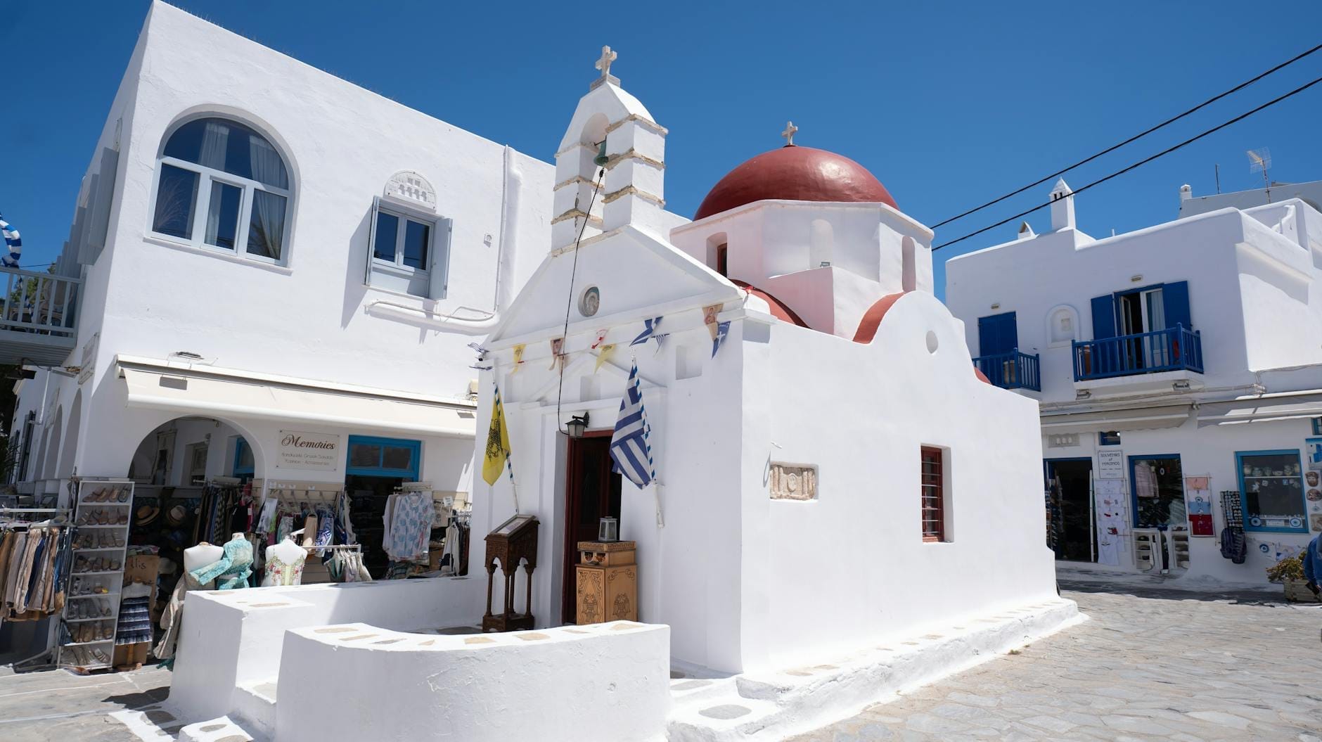 Pequeña iglesia encalada con cúpula roja, banderas griegas y tiendas cercanas en una tranquila calle de Mykonos bajo un cielo azul despejado.