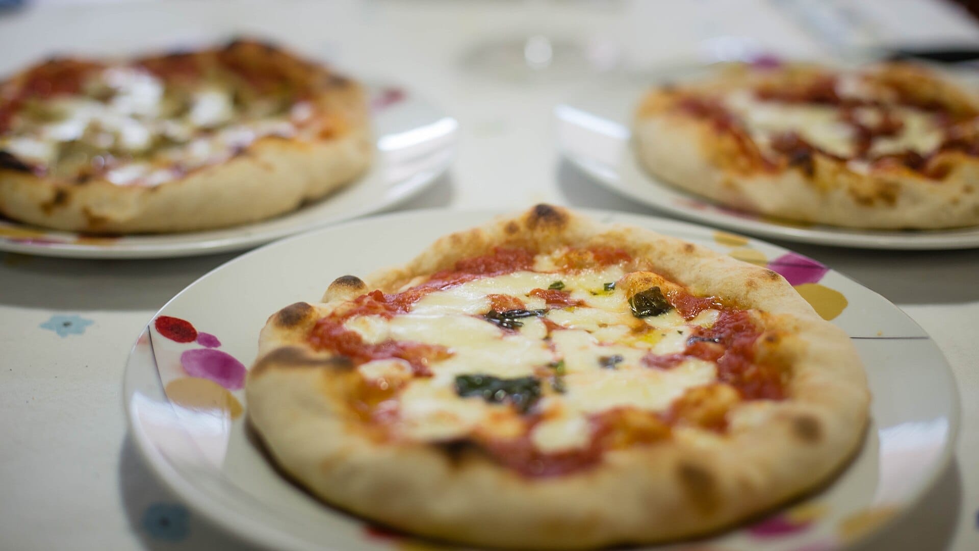 Três pizzas ao estilo napolitano com massa chamuscada, molho de tomate e mozzarella derretida em pratos sobre uma mesa.