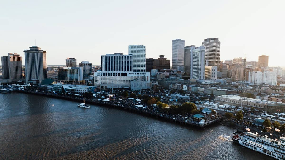 Aerial view of the New Orleans riverfront with city skyline, boats on the Mississippi River, and people along the promenade.